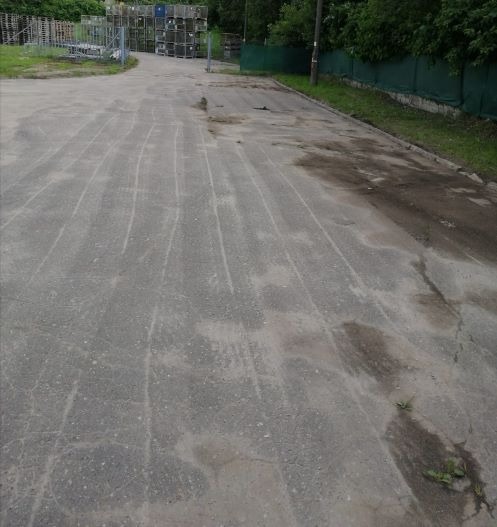 Asfaltowa droga z widocznymi śladami po odśnieżaniu, fragmentami błota i kałużami, w tle magazynowe pojemniki.