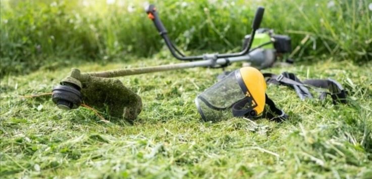 Spalinowa kosa do trawy i żółta osłona twarzy leżą na świeżo skoszonej trawie w słoneczny dzień.