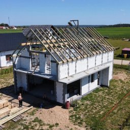 Usługi ogólnobudowlane Adaś - Wznoszenie domu z białych bloczków, widoczne drewniane krokwie dachu, w tle zielone pola i zabudowania, ujęcie z drona.