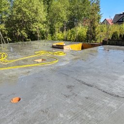 Usługi ogólnobudowlane Adaś - Świeżo wylany, mokry fundament z widocznymi żółtymi elementami szalunku i żółtym kablem na powierzchni. W tle zieleń drzew i zabudowania.