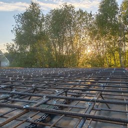 Usługi ogólnobudowlane Adaś - Zbrojenie płyty fundamentowej domu jednorodzinnego w promieniach zachodzącego słońca, widok z bliska na siatkę stalową z elementami dystansowymi.