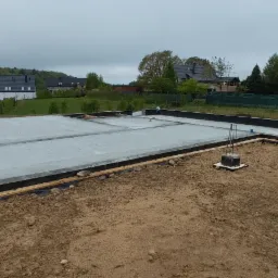 Zalana płyta fundamentowa z wystającymi prętami zbrojeniowymi, otoczona szalunkiem. W tle zabudowa jednorodzinna i zieleń. Etap budowy domu.