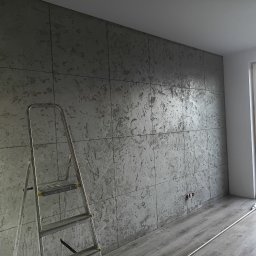 Usługi ogólnobudowlane Adaś - Ściana wykończona szarym tynkiem dekoracyjnym imitującym beton, podzielona na kwadraty. Aluminiowa drabina oparta o ścianę, widoczne gniazdka elektryczne i fragment podłogi.