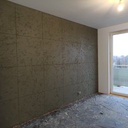 Usługi ogólnobudowlane Adaś - Ściana w pokoju wykończona dekoracyjnym tynkiem strukturalnym w odcieniach szarości, podzielona na kwadratowe panele. Podłoga zabezpieczona folią malarską, widoczne okno balkonowe.