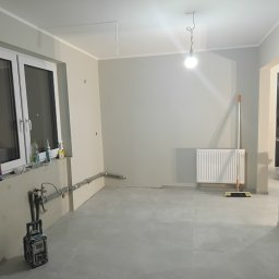 Usługi ogólnobudowlane Adaś - Szare płytki podłogowe w nowym, jasnym pomieszczeniu z grzejnikiem, oknem i narzędziami. Widoczne rury i niedokończone wykończenia.