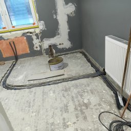 Usługi ogólnobudowlane Adaś - Szare rury ogrzewania podłogowego ułożone w pętli na przygotowanym podłożu, widoczny grzejnik, ściany w szarym kolorze w trakcie prac wykończeniowych, okno z folią ochronną.