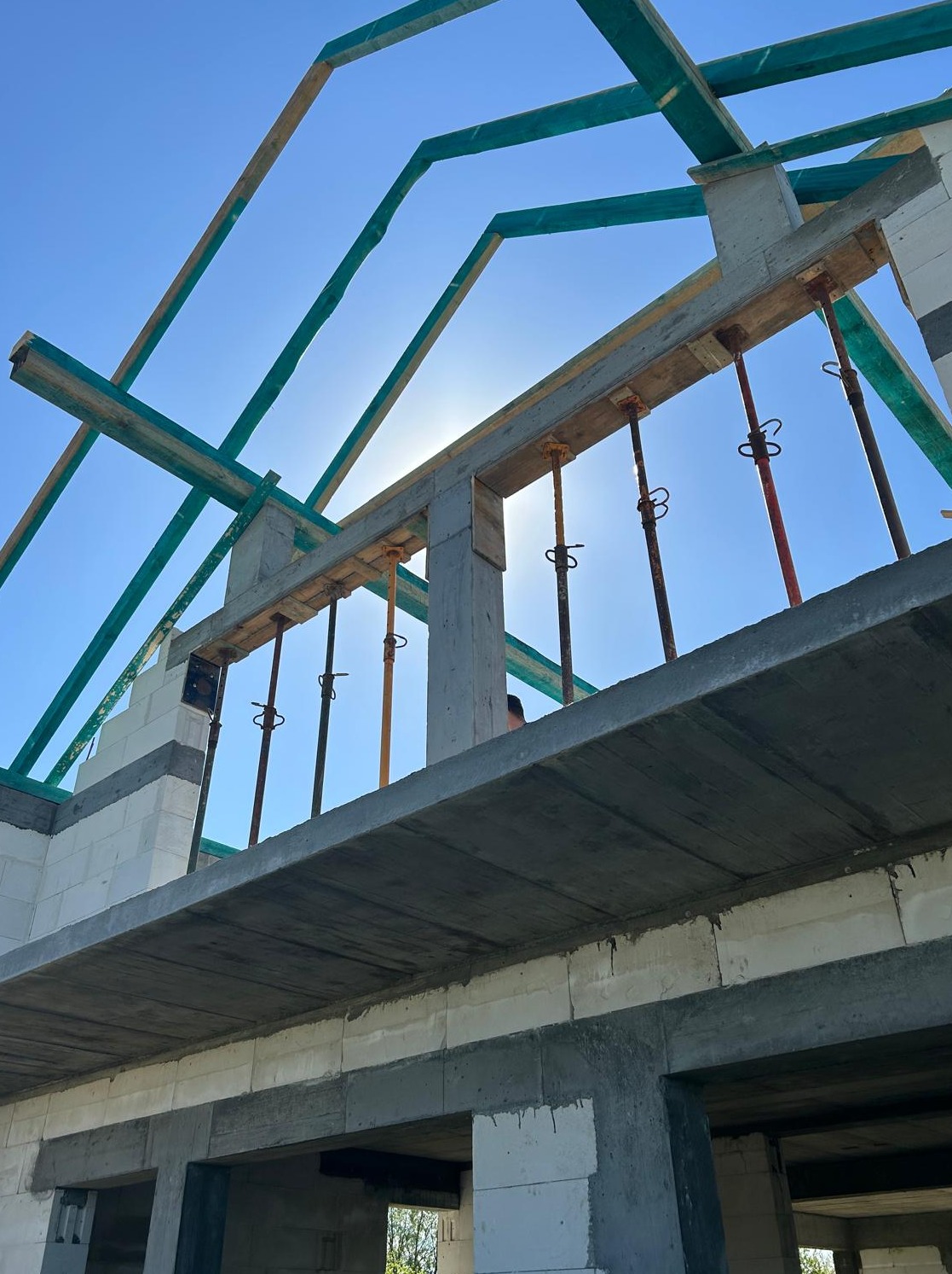 Wznoszenie domu z betonu komórkowego: widok na konstrukcję stropu i zbrojenie, podpory stalowe podtrzymujące szalunek, fragment więźby dachowej w tle.