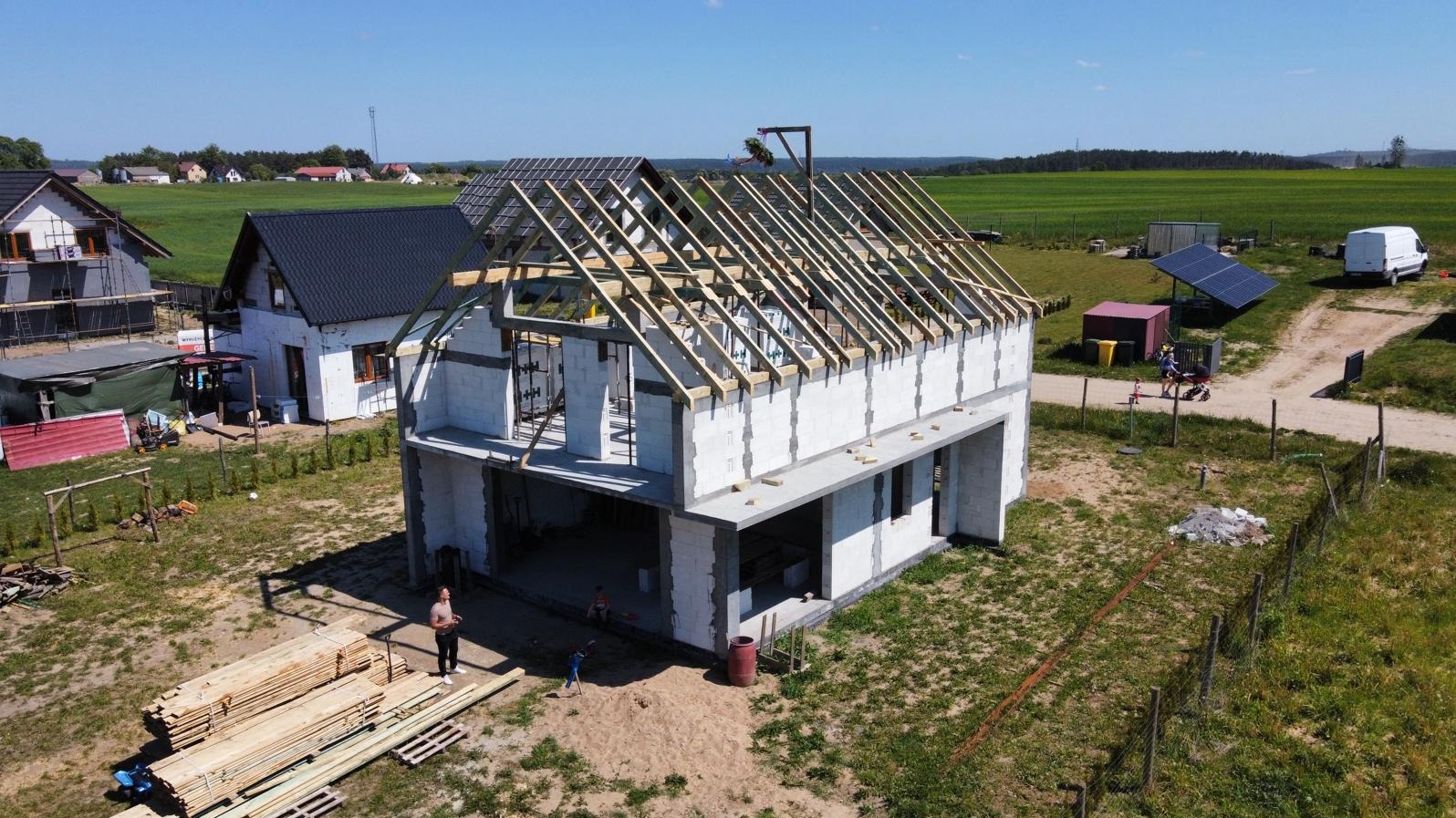 Wznoszenie domu z białych bloczków, widoczne drewniane krokwie dachu, w tle zielone pola i zabudowania, ujęcie z drona.