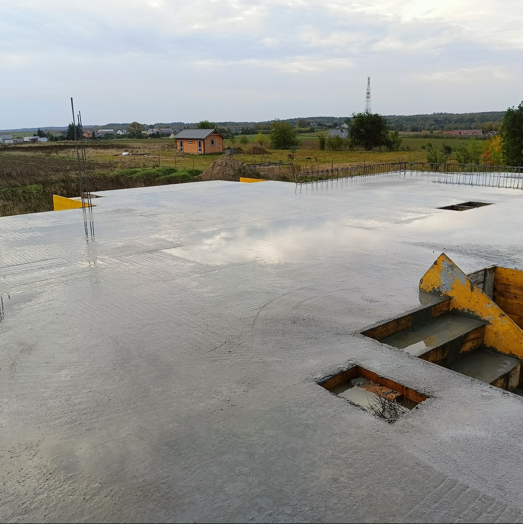 Świeżo wylana, mokra płyta fundamentowa z widocznymi zbrojeniami i otworami, żółte szalunki krawędziowe, w tle krajobraz wiejski i mały domek.