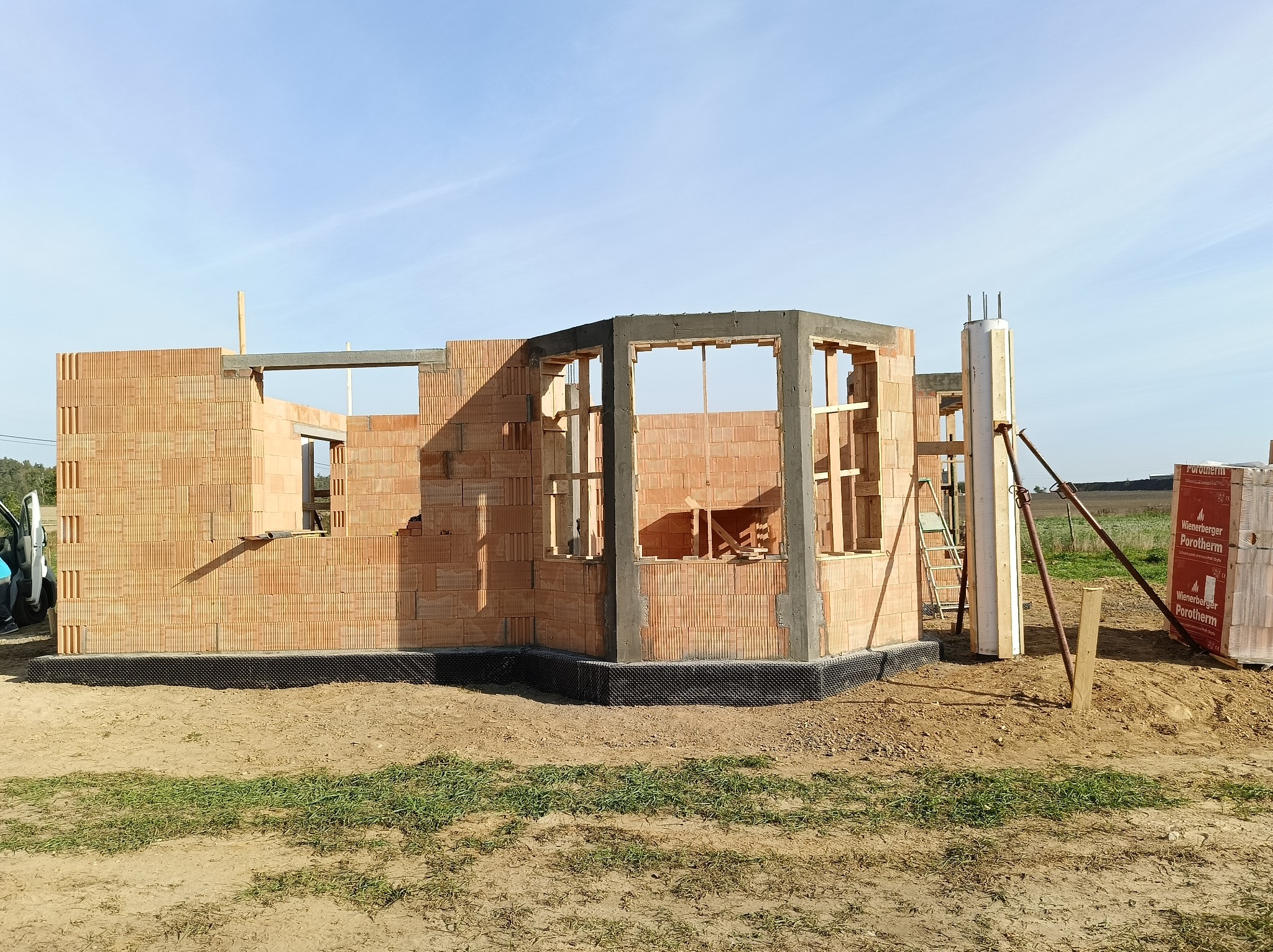 Wznoszenie ścian domu z pustaków ceramicznych Porotherm, widoczne zbrojenie nadproży i słupów, teren budowy z widoczną hydroizolacją fundamentów.