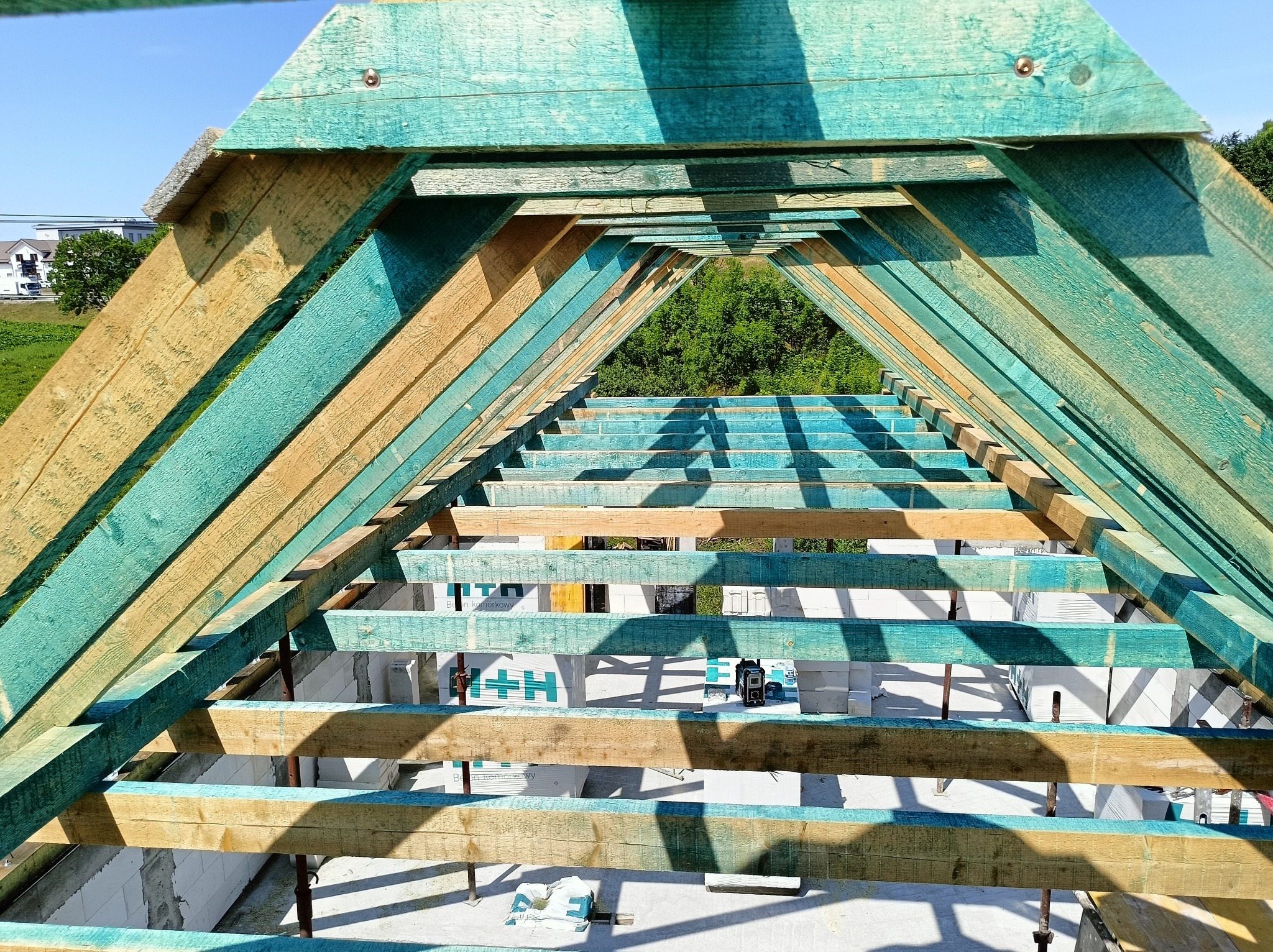 Wnętrze konstrukcji dachu z belek pomalowanych na zielono i naturalne drewno, widok z dołu na niebo i drzewa, w tle białe bloczki z betonu komórkowego.