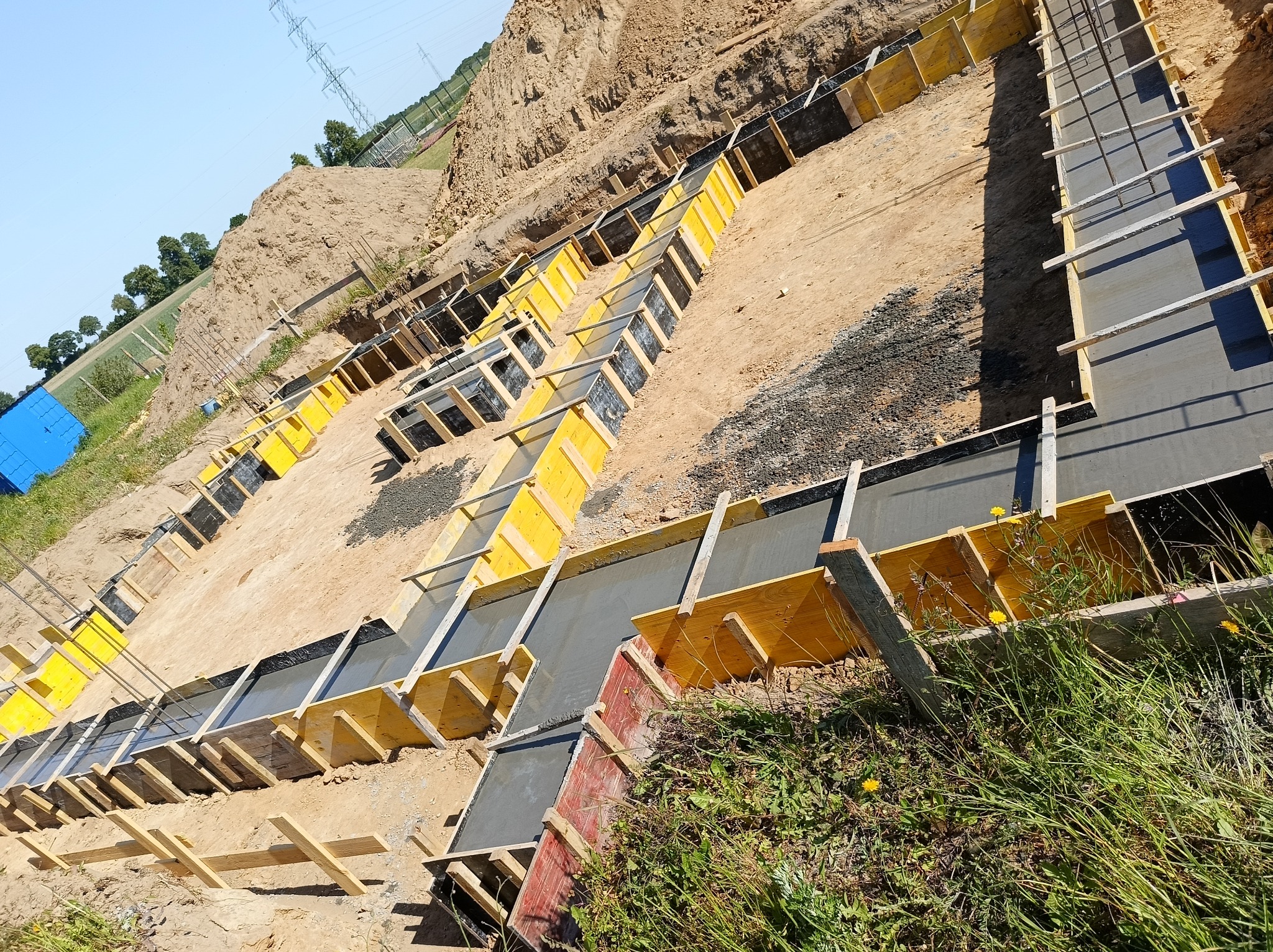 Wykop pod fundamenty z żółtym szalunkiem wypełniony świeżym betonem, widok z góry na tle ziemi i zieleni.