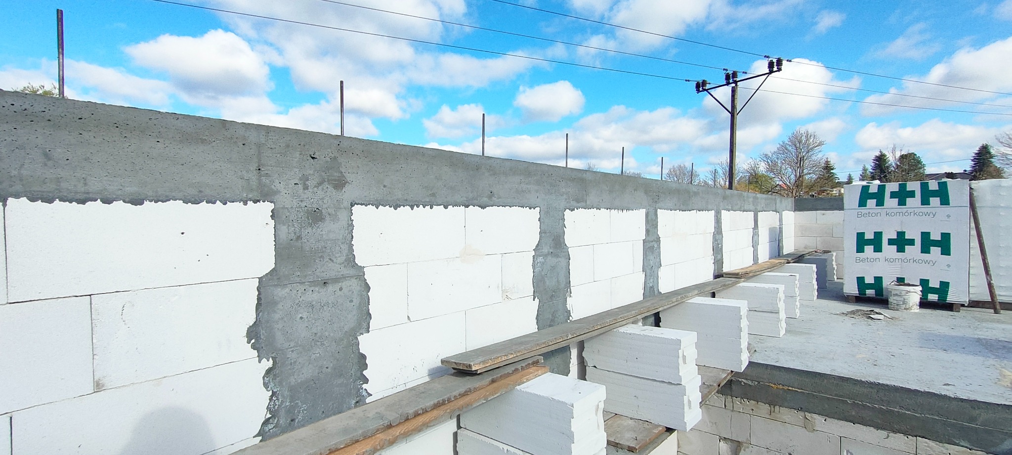 Wznoszenie ściany z białych bloczków betonowych H+H na fundamencie, widoczne zbrojenia pionowe i paczki bloczków na paletach w tle, słup energetyczny z przewodami na niebie.