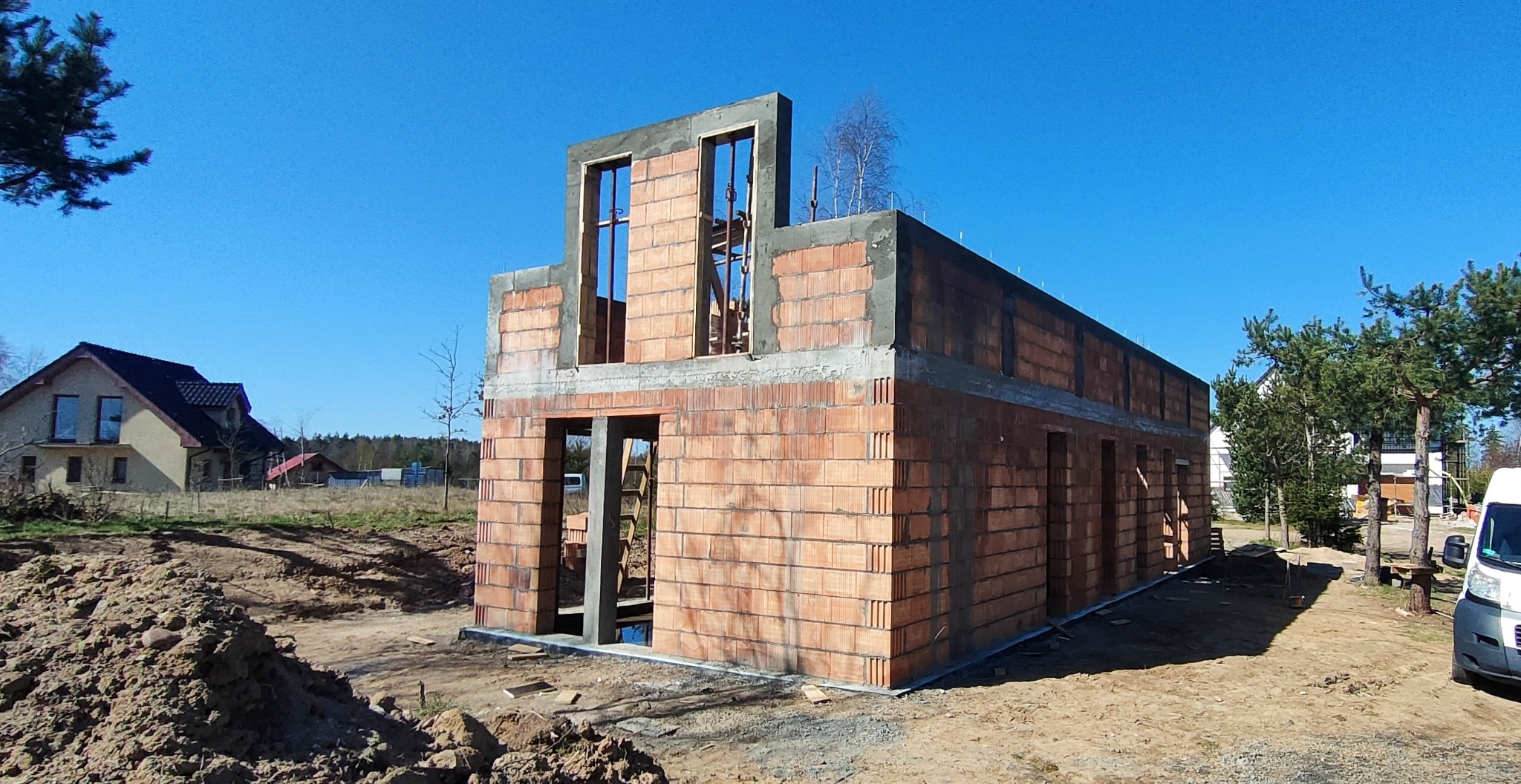 Surowy stan domu z czerwonej cegły, widoczne otwory okienne i drzwiowe, zarys drugiego piętra, na tle błękitnego nieba i częściowo zalesionego terenu.