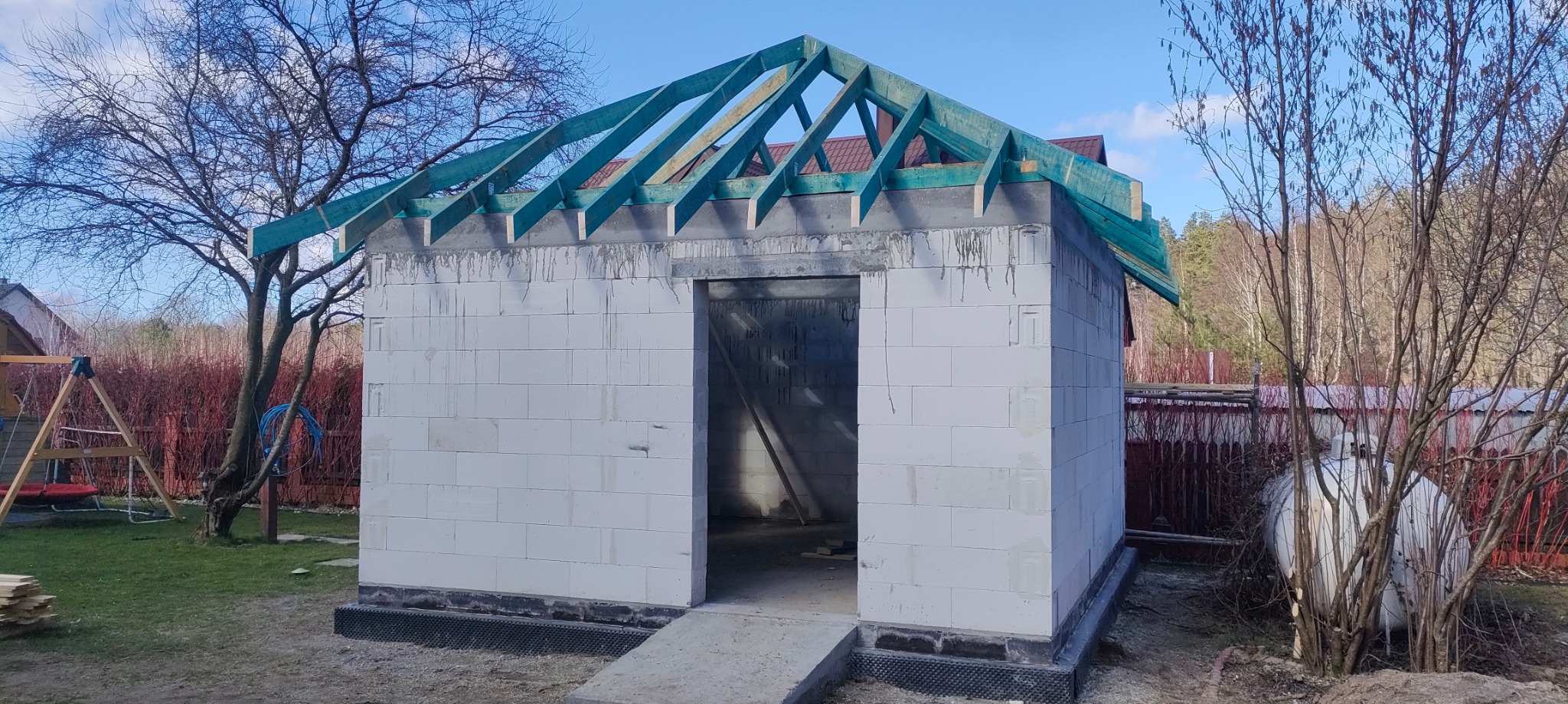 Surowy budynek z białych bloczków z zieloną konstrukcją dachu w trakcie budowy, widoczny wjazd i otoczenie działki z drzewami.