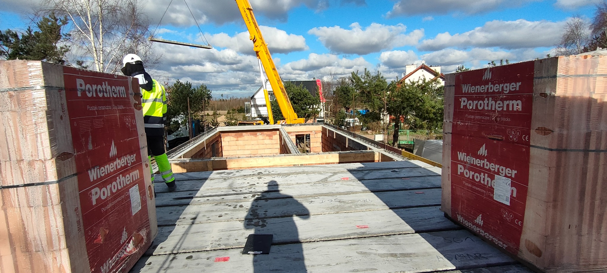 Budowa domu: widok z góry na wznoszone ściany z czerwonej cegły Porotherm, z dźwigiem w tle i pracownikiem w odblaskowej kamizelce, palety z cegłami na pierwszym planie.