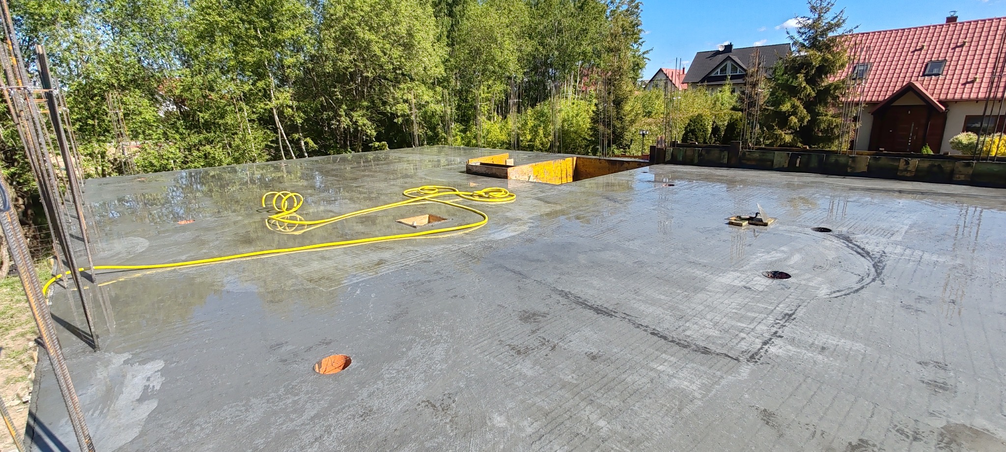 Świeżo wylany, mokry fundament z widocznymi żółtymi elementami szalunku i żółtym kablem na powierzchni. W tle zieleń drzew i zabudowania.