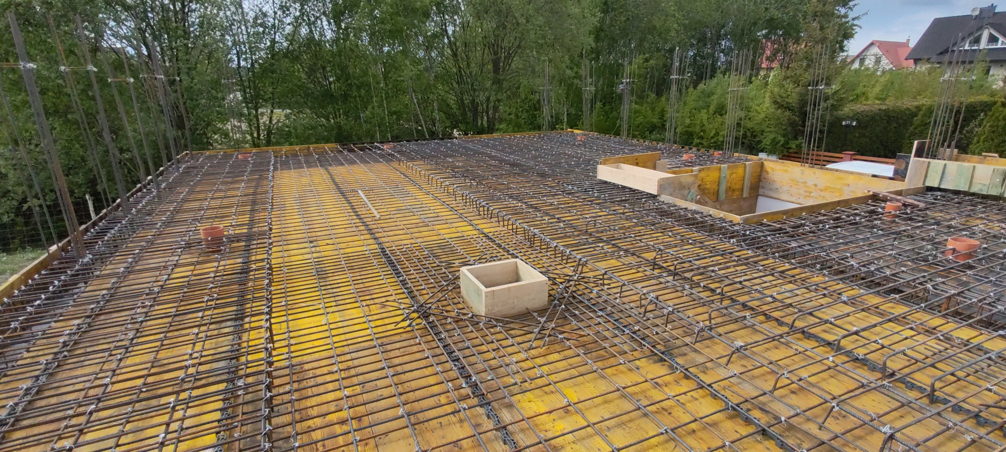 Zbrojenie stropu budynku z żółtymi szalunkami i elementami drewnianymi, widok z góry na tle zieleni i zabudowań. Układ prętów tworzy regularną siatkę.