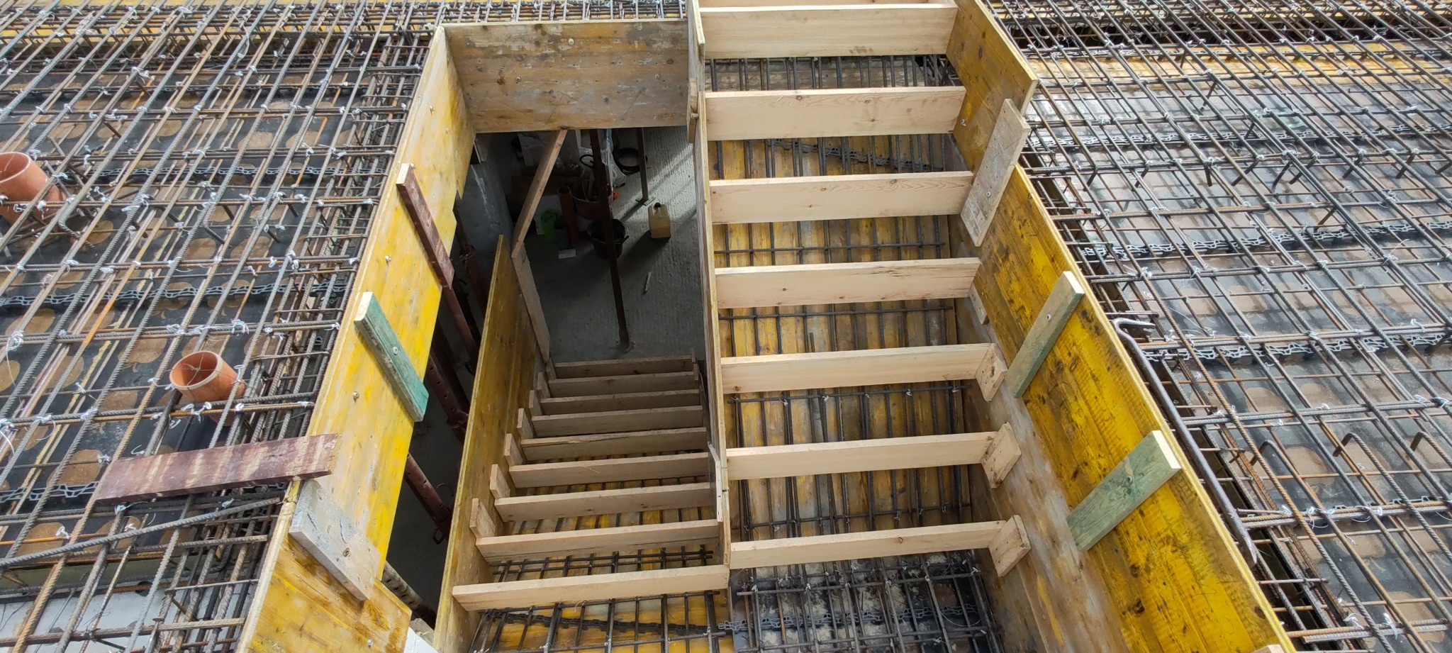 Zbrojenie stropu z drewnianym szalunkiem schodów. Widok z góry na siatkę zbrojeniową i deskowanie w kolorze żółtym, przygotowane do wylania betonu.