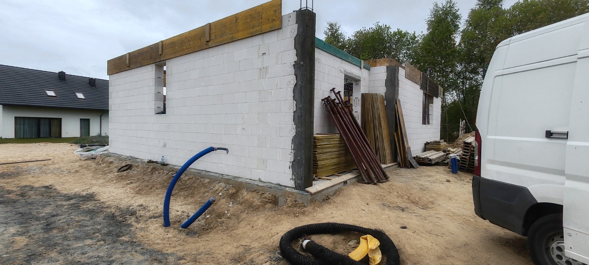 Budowa domu z białych bloczków: ściany w trakcie murowania, widoczne podpory i narzędzia. W tle dom z ciemnym dachem i biała furgonetka.