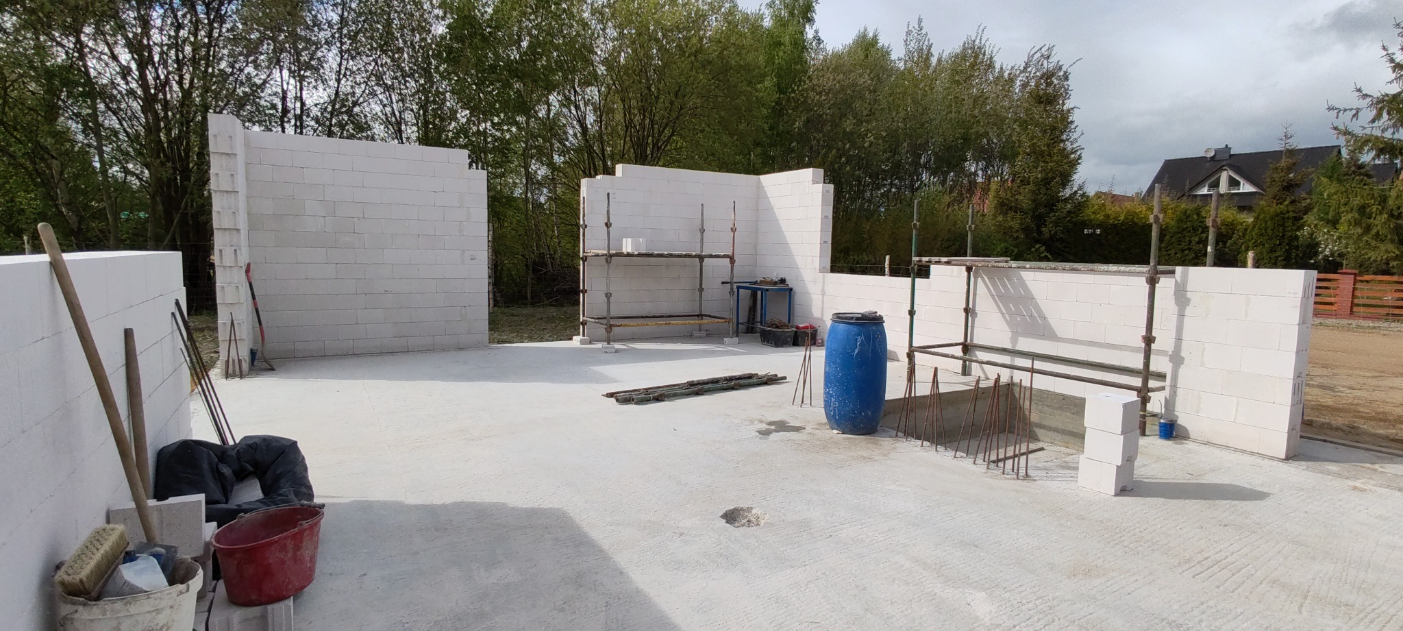 Budowa domu z białych bloczków: ściany w trakcie wznoszenia, widoczne rusztowania, narzędzia i niebieska beczka. Etap budowy w słoneczny dzień.