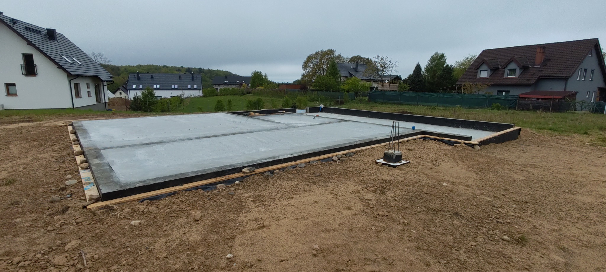 Zalana płyta fundamentowa z wystającymi prętami zbrojeniowymi, otoczona szalunkiem. W tle zabudowa jednorodzinna i zieleń. Etap budowy domu.