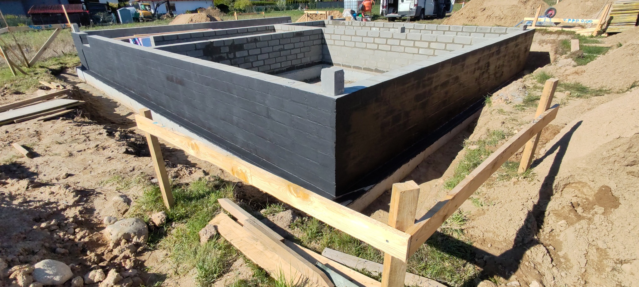 Zabezpieczone, pomalowane na czarno fundamenty z bloczków betonowych w wykopie, widoczne drewniane podpory szalunku, w tle sprzęt budowlany.