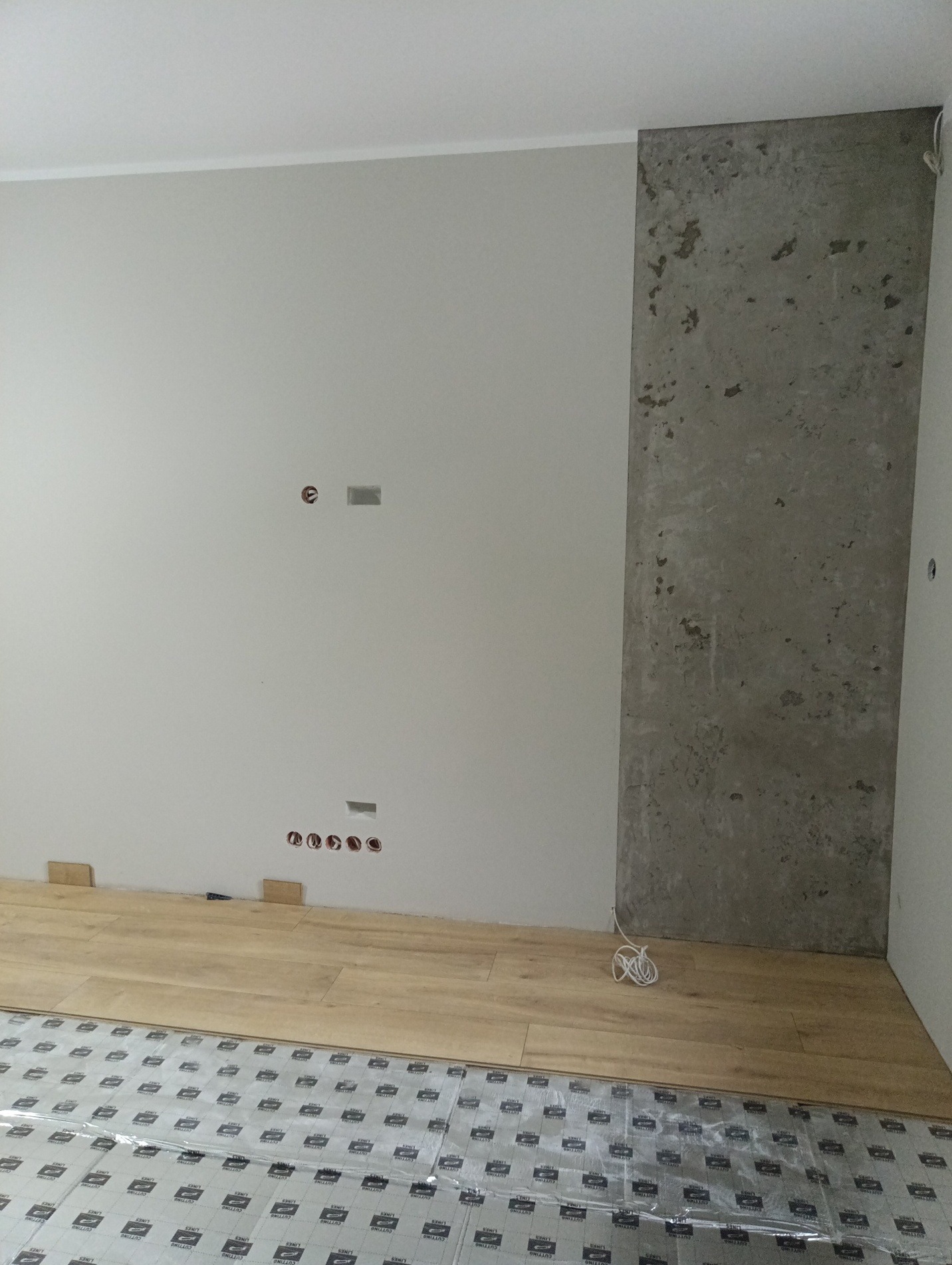 Wnętrze w trakcie wykańczania: częściowo ułożona drewniana podłoga na folii ochronnej, fragment ściany z betonową płytą dekoracyjną i przygotowane gniazdka elektryczne.
