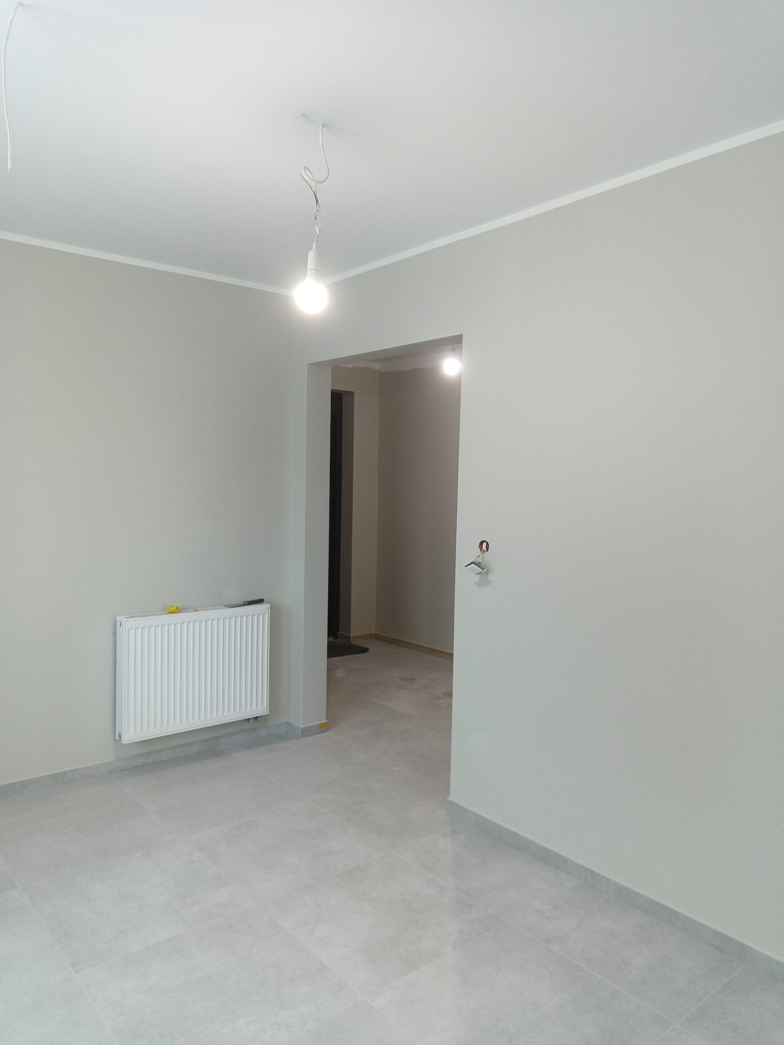 Jasne, świeżo wykończone wnętrze z szarymi ścianami, płytkami podłogowymi i białym grzejnikiem. Widoczne gniazdko elektryczne i żarówka na kablu.