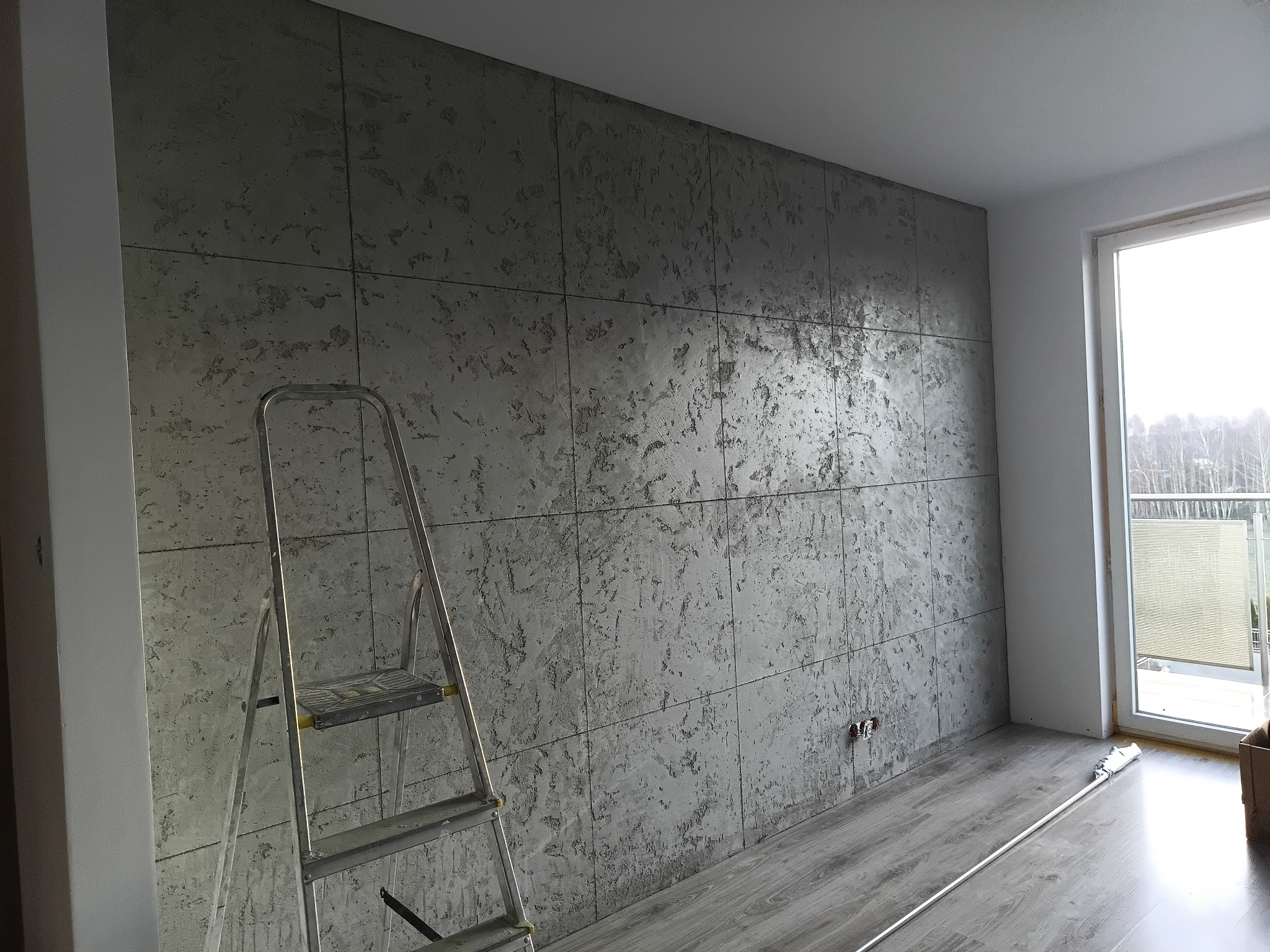 Ściana wykończona szarym tynkiem dekoracyjnym imitującym beton, podzielona na kwadraty. Aluminiowa drabina oparta o ścianę, widoczne gniazdka elektryczne i fragment podłogi.
