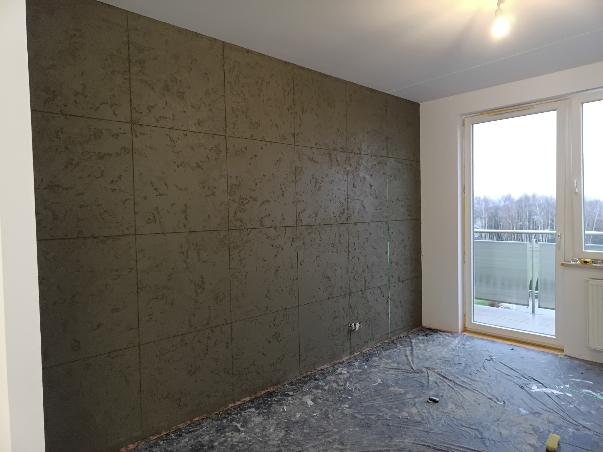 Ściana w pokoju wykończona dekoracyjnym tynkiem strukturalnym w odcieniach szarości, podzielona na kwadratowe panele. Podłoga zabezpieczona folią malarską, widoczne okno balkonowe.