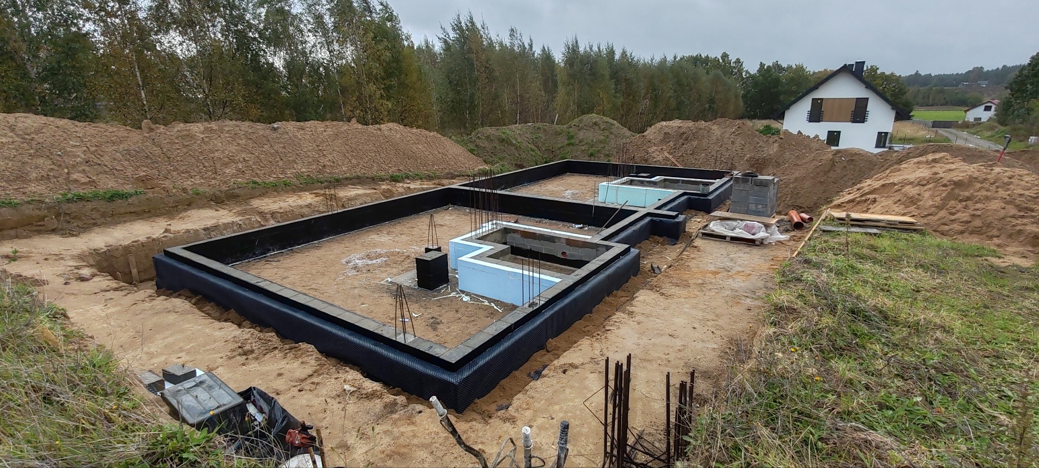 Zabezpieczony fundament domu jednorodzinnego z hydroizolacją i ociepleniem, widok z góry na wykop z ziemią, w tle dom w budowie i drzewa.