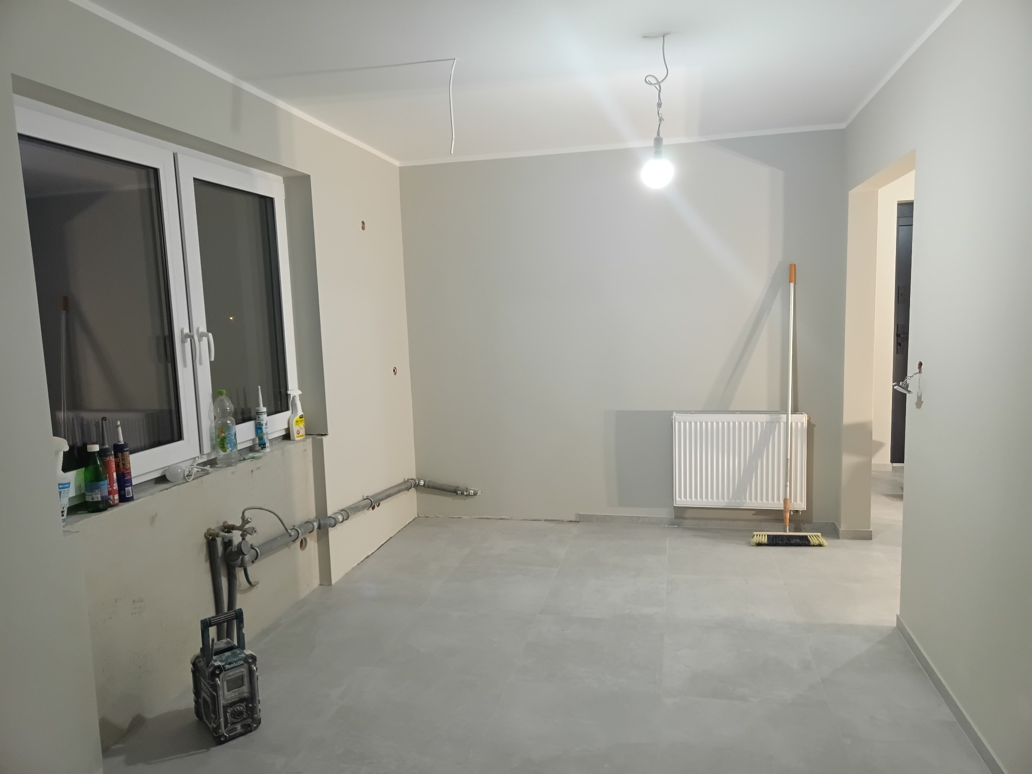 Szare płytki podłogowe w nowym, jasnym pomieszczeniu z grzejnikiem, oknem i narzędziami. Widoczne rury i niedokończone wykończenia.
