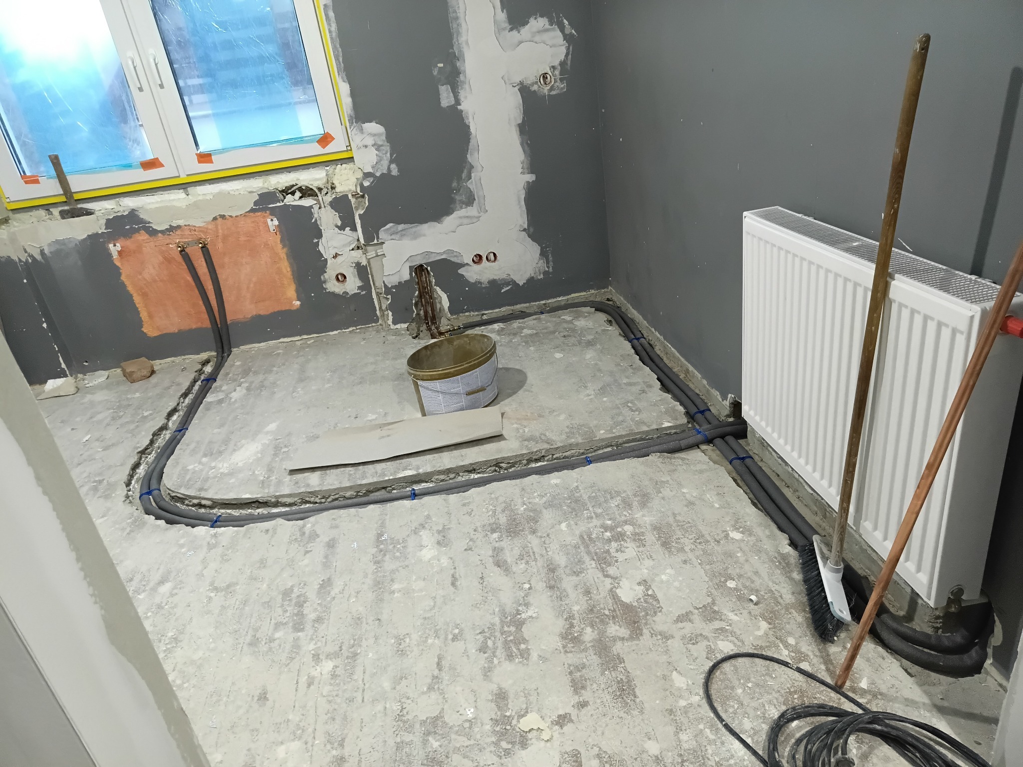 Szare rury ogrzewania podłogowego ułożone w pętli na przygotowanym podłożu, widoczny grzejnik, ściany w szarym kolorze w trakcie prac wykończeniowych, okno z folią ochronną.