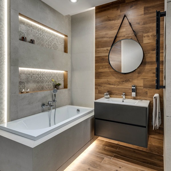 Nowoczesna łazienka z drewnopodobnymi płytkami na ścianie, okrągłym lustrem na pasku i podświetlanymi wnękami z dekoracyjnymi płytkami nad wanną.