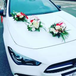 Biały samochód udekorowany trzema bukietami z białych kwiatów i czerwonymi wstążkami na masce, przygotowany na uroczystość.