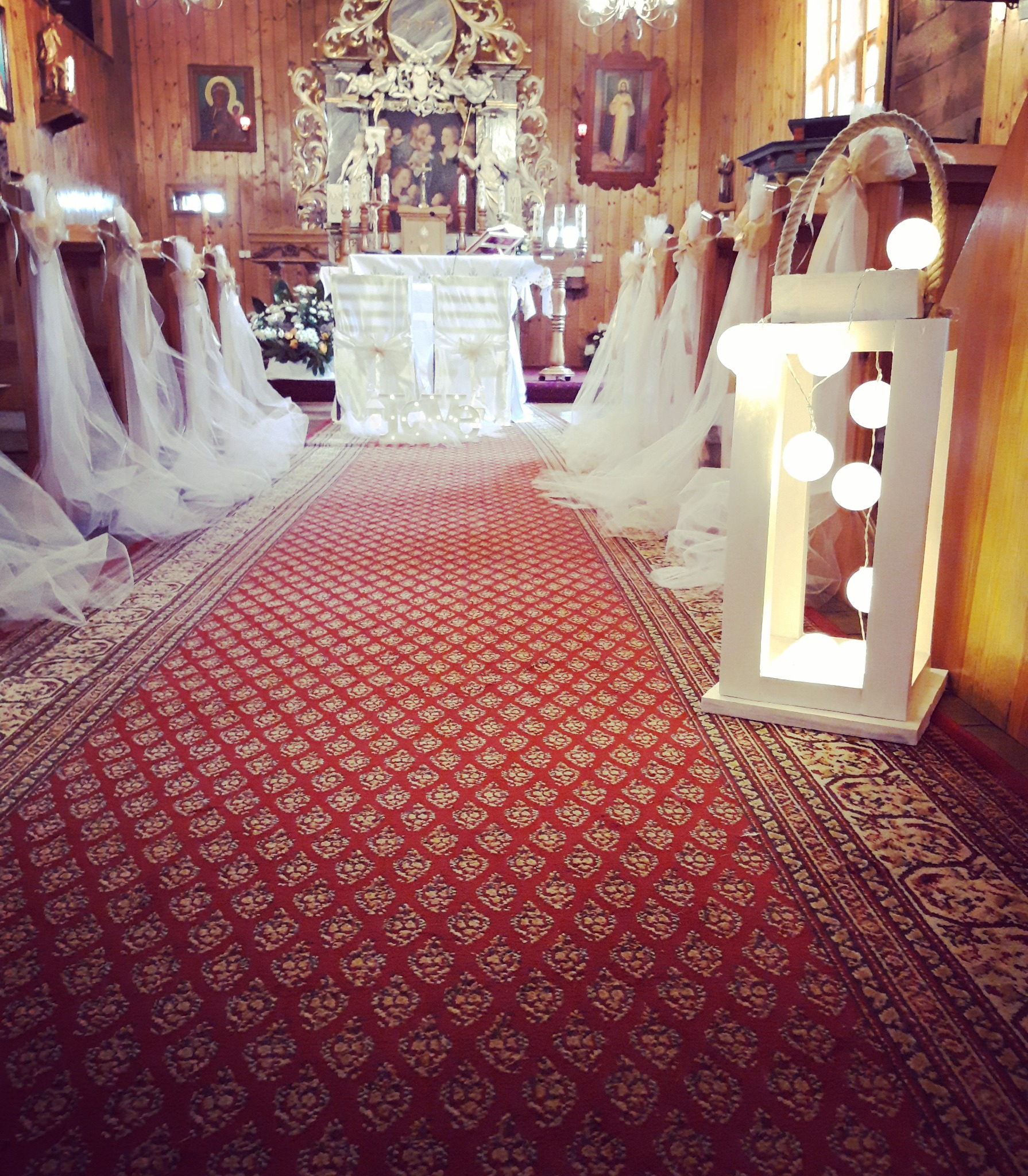 Udekorowane wnętrze kościoła na uroczystość ślubną: czerwony dywan, białe dekoracje na ławkach i oświetlony lampion.