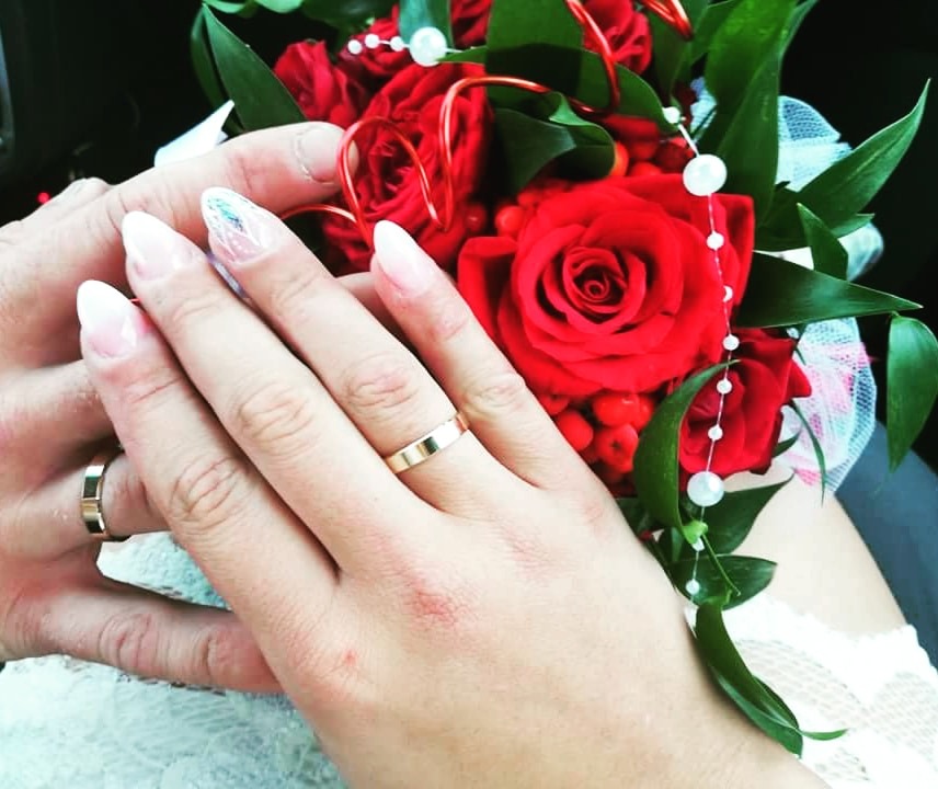 Dłonie pary młodej z obrączkami na tle bukietu czerwonych róż z białymi perłami.