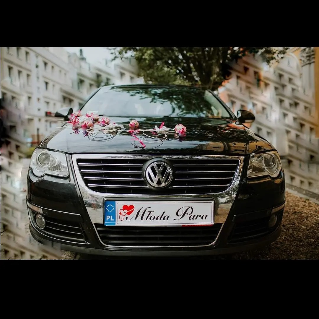 Czarny Volkswagen Passat z dekoracją z różowych kwiatów i wstążek na masce, tablica rejestracyjna z napisem 'Młoda Para' na tle miejskiej zabudowy.