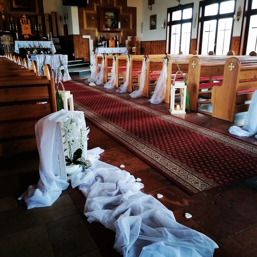 Dekoracja kościoła na ślub: białe orchidee w drewnianej skrzynce, tiul na podłodze, czerwony dywan i lampiony przy ławkach.