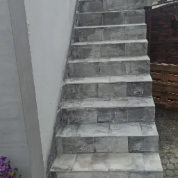 Zewnętrzne schody wyłożone szarą, prostokątną płytką imitującą kamień, z widocznymi śladami fugowania, prowadzące do budynku z szarą elewacją i kwiatami w donicy obok.