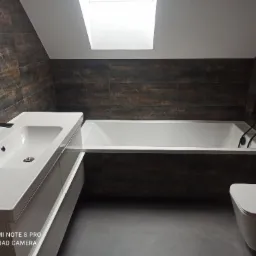 Nowoczesna łazienka na poddaszu z brązowymi płytkami imitującymi drewno, białą wanną i umywalką z czarną armaturą oraz oknem dachowym.