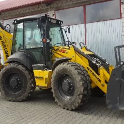 Żółto-czarna koparko-ładowarka New Holland 115C zaparkowana na kostce brukowej przed budynkiem z blachy falistej i cegły, z widocznymi łyżkami z przodu i z tyłu.