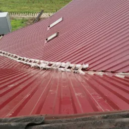 Czerwony dach z blachy trapezowej z zamontowanymi oknami dachowymi i zabezpieczoną drabiną dekarską, widok z góry podczas prac dekarskich.