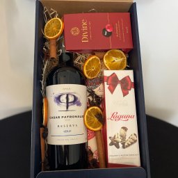 RAVEL POLAND Spółka Cywilna - Elegancki kosz prezentowy: wino Casas Patronales Merlot, czekoladki Divine i Laguna, dekoracje z pomarańczy i cynamonu, całość w granatowym pudełku.