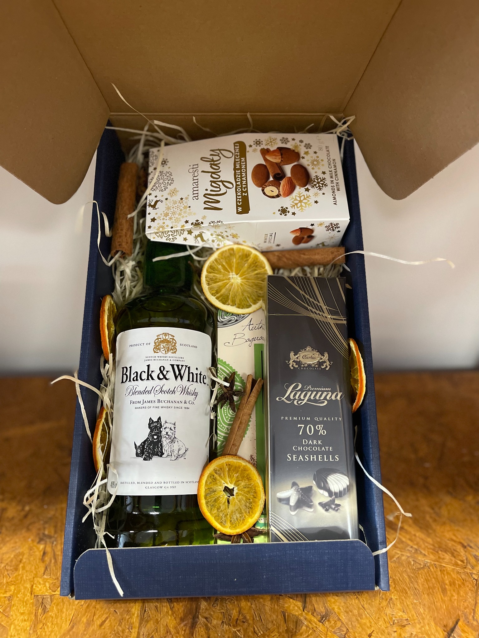 Elegancki kosz prezentowy: whisky Black&White, czekolada Laguna, migdały w czekoladzie, dekoracje z pomarańczy i cynamonu w granatowym pudełku.