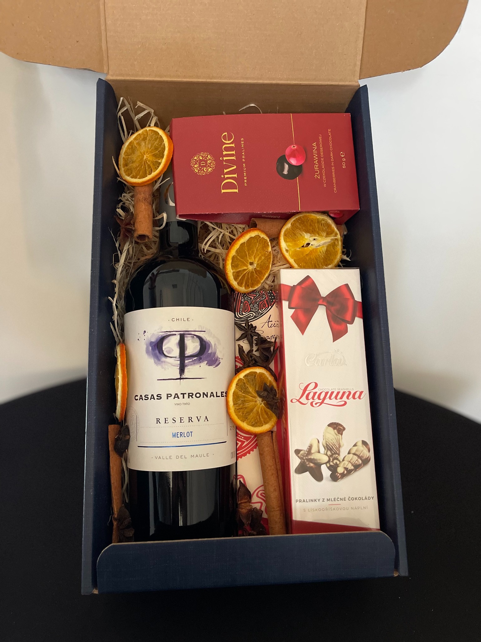 Elegancki kosz prezentowy: wino Casas Patronales Merlot, czekoladki Divine i Laguna, dekoracje z pomarańczy i cynamonu, całość w granatowym pudełku.