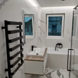 Nowoczesna łazienka z marmurowymi płytkami, czarną armaturą i grzejnikiem drabinkowym, odbicie budynku w oknie i lustrze.