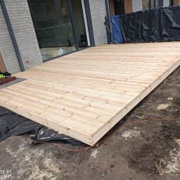 Klaudia Kajtarek - Drewniany taras w trakcie budowy, widoczne deski tworzące powierzchnię, czarna folia pod spodem, narzędzia i materiały budowlane obok, ziemia wokół.