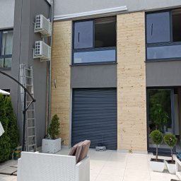 Klaudia Kajtarek - Elewacja budynku z drewnianą okładziną, widoczne okna, brama garażowa, klimatyzatory na szarej ścianie, drabina oparta o ścianę, białe meble ogrodowe na jasnym tarasie.